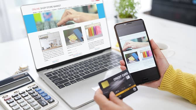Quasiment toutes les activités en ligne peuvent contribuer à votre empreinte numérique. L’image montre quelqu’un tenir son téléphone et sa carte bancaire devant un écran d’ordinateur, avec une fenêtre ouverte sur une boutique en ligne.